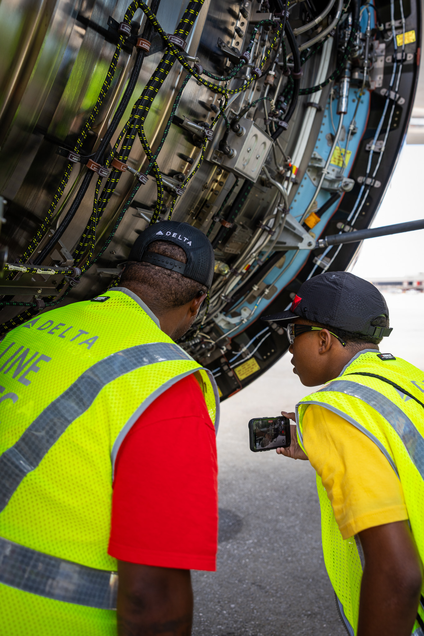A student and aviation professional examine exposed aircraft engine components while the student records details on a smartphone.