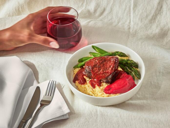 Beef short rib meal in white bowl, with woman's hand gripping glass of red wine to the left of the dish.