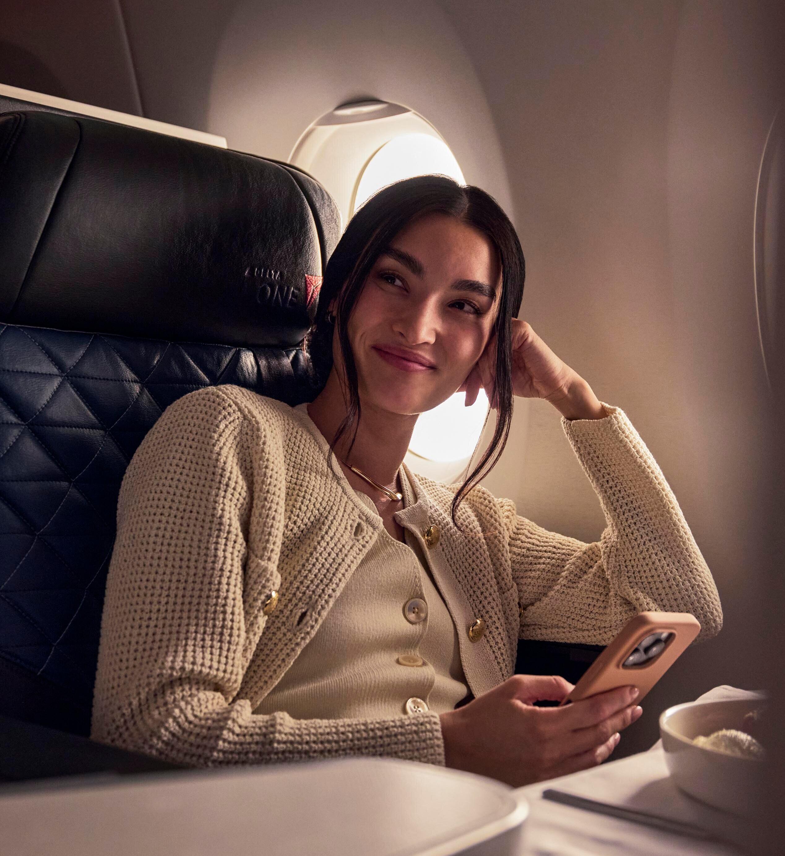 Person seated in a premium airplane cabin by a window, holding a smartphone while relaxing in a spacious seat.