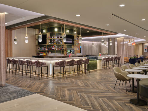 Lounge bar with high‑back stools, illuminated shelving, warm lighting and adjacent seating areas with small tables