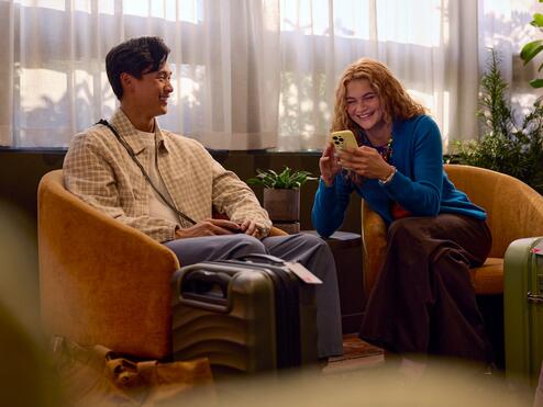 Two travelers sit in lounge chairs with rolling suitcases nearby, one looking at a smartphone, in a softly lit indoor waiting area with plants and sheer curtains.
