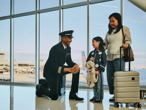 Delta pilot kneeling beside a young child holding a stuffed animal at an airport gate, while an adult stands nearby with rolling luggage; airplane visible through large windows in the background.