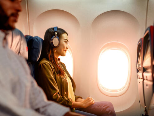 Delta customer on board an airplane wearing headphones sitting by a window, with warm sunlight illuminating the cabin and seatback screens visible.