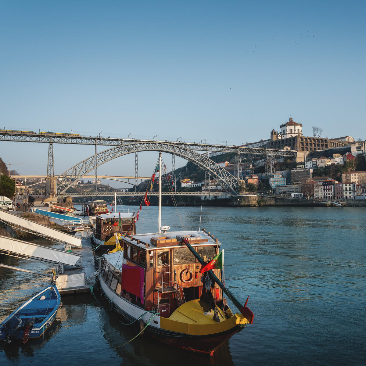 Colorful boats on a river with a large metal arch bridge and hillside buildings in the background.