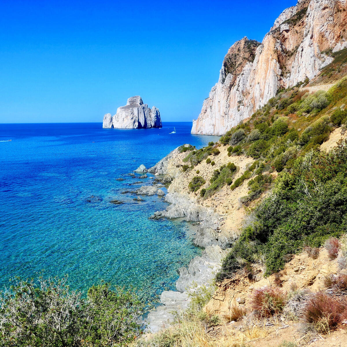 Clear turquoise sea beside rugged cliffs with a large rock formation offshore.