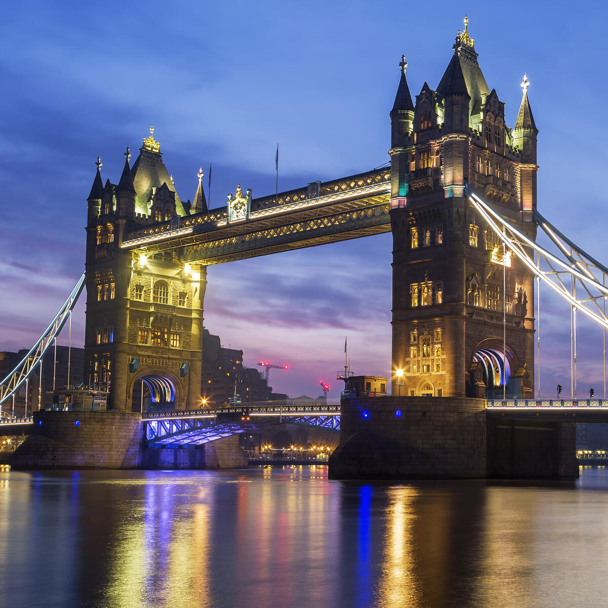Tower Bridge illuminated at dusk, with glowing lights reflecting across the calm river and a blue‑purple sky creating a dramatic backdrop over the historic structure.