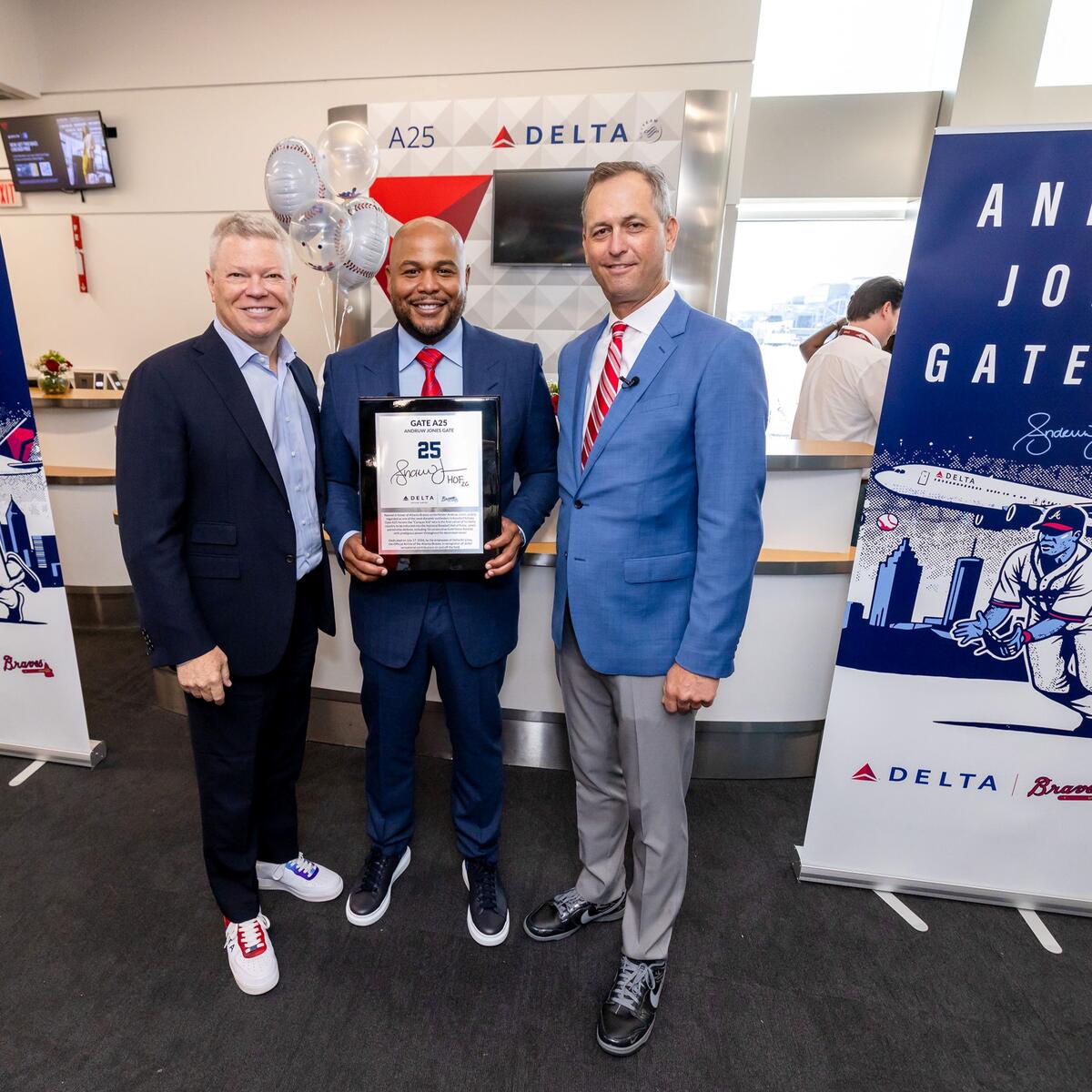 Three people pose at a Delta airport gate ceremony, with the center person holding a commemorative plaque. Banners on both sides read “Andruw Jones Gate A25,” featuring baseball-themed artwork and Delta branding.