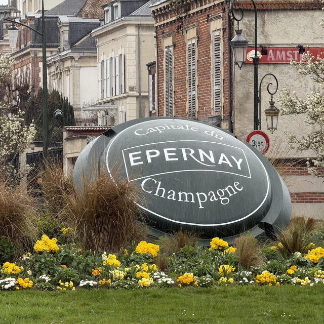 A large, green, shield-shaped monument with the white text "Capitale du Champagne, EPERNAY" sits in the middle of a floral roundabout, surrounded by yellow flowers and classic French buildings under a cloudy sky.