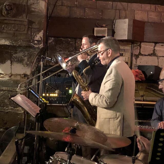 Two musicians play trombone and saxophone during an intimate jazz set in a dimly lit stone-walled venue.