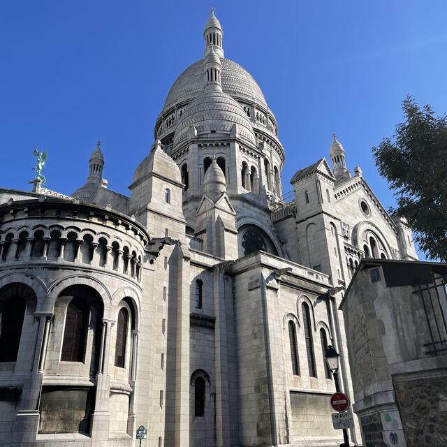 The large, white stone Sacré-Cœur Basilica in Paris stands under a clear blue sky, with its central dome and arcaded apse prominent, and people walking below.