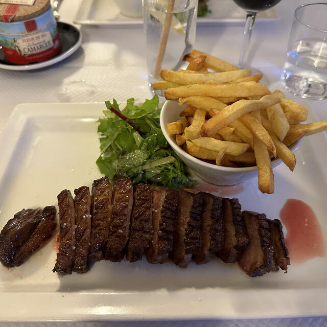 A white rectangular plate holds sliced grilled meat, a small bowl of golden french fries, and a side salad, set on a table with salt and water glasses.