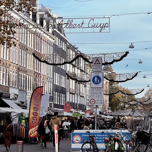 A busy outdoor market street lined with tall brick buildings, vendor stalls, signage, and hanging decorations, with crowds walking beneath an overhead sign that reads ‘Albert Cuyp.’