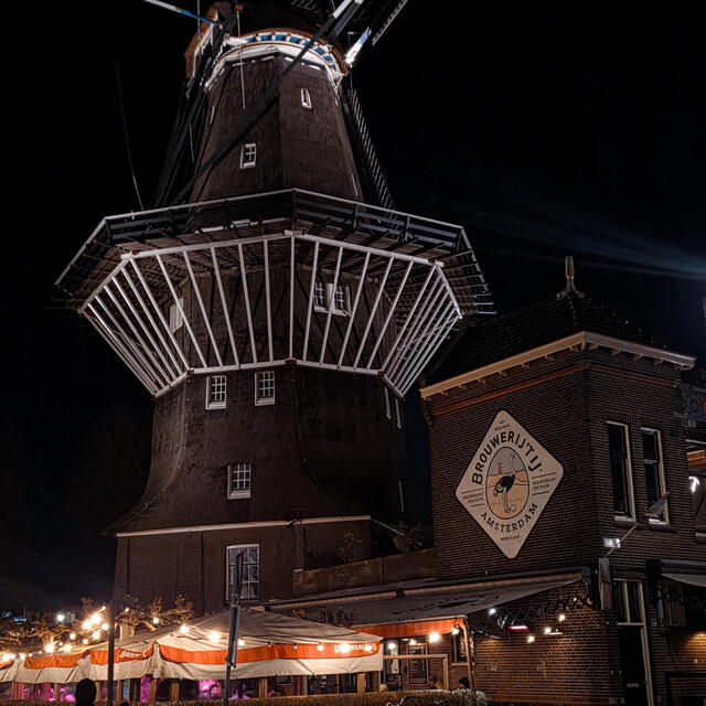 A large traditional windmill and adjacent brick brewery building are illuminated at night, with outdoor seating and lights beneath.