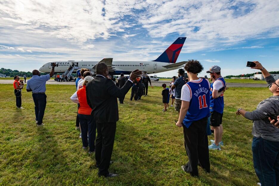 Spectators stand on grass watching a Delta airplane taxi on the runway at an airport.