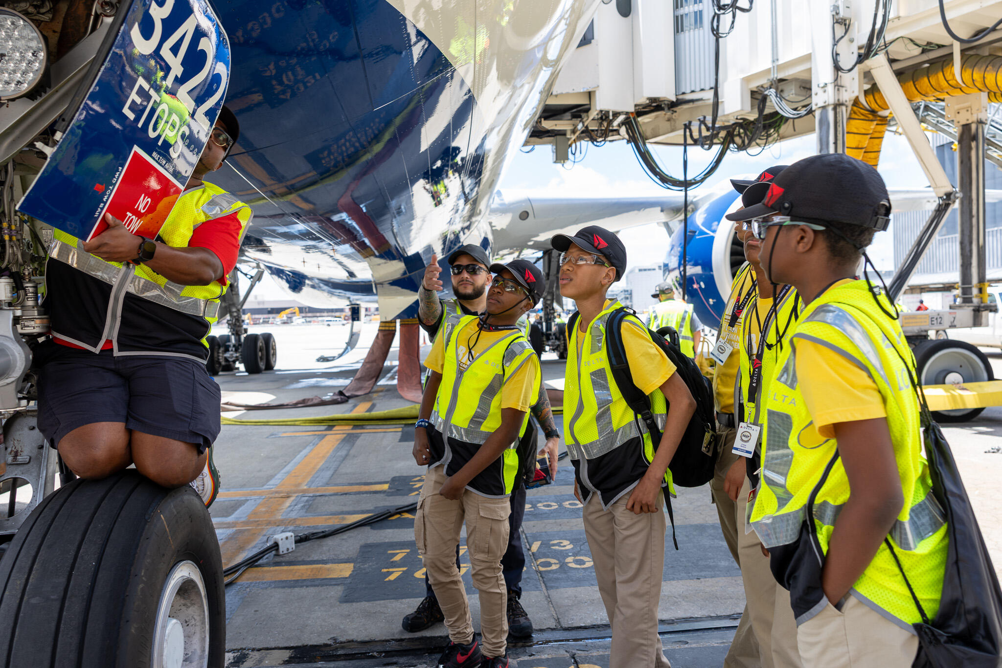Students in safety vests gather beneath a Delta aircraft as an aviation professional points out features near the landing gear on the airport ramp.