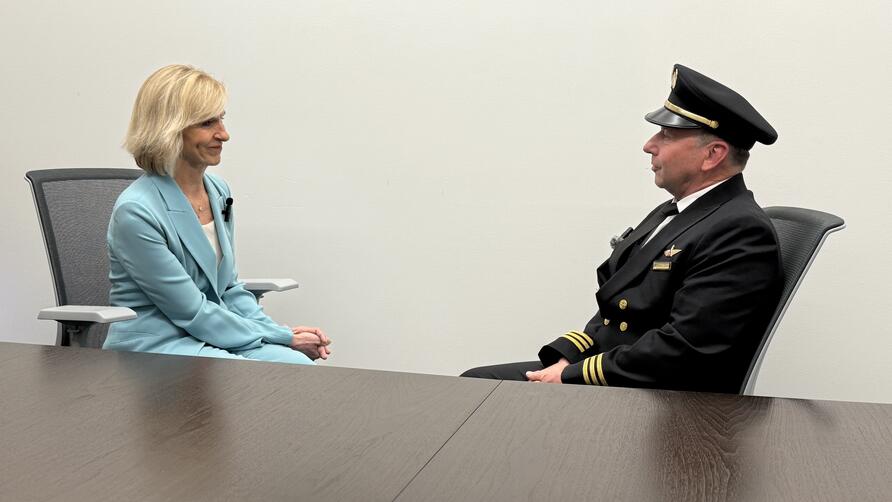 Allison Ausband and Tom Cohan sit facing each other at a conference table, Allison in a business suit and Tom in a pilot uniform.