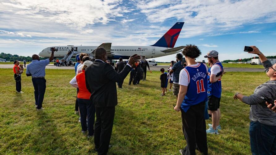 Spectators stand on grass watching a Delta airplane taxi on the runway at an airport.