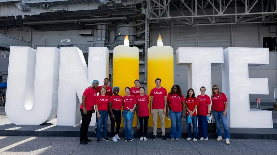 Delta volunteers in red shirts stand in front of a large “UNITE” display with two candle-shaped letters, commemorating 9/11 aboard an aircraft carrier.