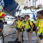 Students in safety vests gather beneath a Delta aircraft as an aviation professional points out features near the landing gear on the airport ramp.
