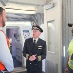 Three airport employees standing and talking near an aircraft boarding door, including a pilot in uniform and two workers wearing high-visibility safety vests.