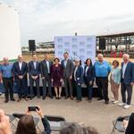Group photo of event attendees standing in front of an industrial biofuels facility, with storage tanks, railcars, and a branded backdrop visible behind them while audience members take photos in the foreground.