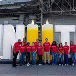 Delta volunteers in red shirts stand in front of a large “UNITE” display with two candle-shaped letters, commemorating 9/11 aboard an aircraft carrier.