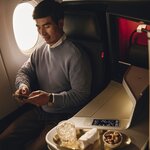 Customer seated in a premium airline suite, using a mobile phone beside a tray with a beverage and snacks.