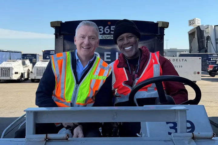 Two workers in high-visibility vests sitting on an airport ground vehicle near Delta equipment and a boarding gate.
