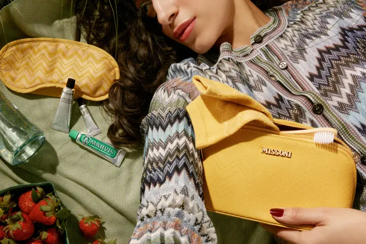 Person lying on grass with a yellow Missoni pouch, eye mask, skincare items, toothpaste, and strawberries arranged on a picnic cloth.