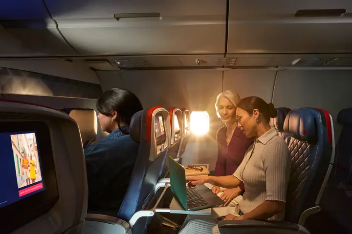 Two female Delta customers on board an aircraft working on laptops, with warm sunlight streaming through the windows.