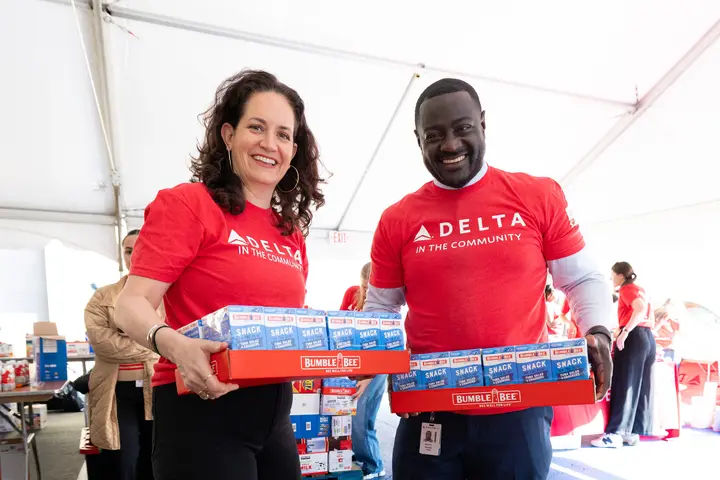 Two Delta volunteers wearing red “Delta in the Community” shirts carry boxes of snack food at a community packing event inside a large tent.