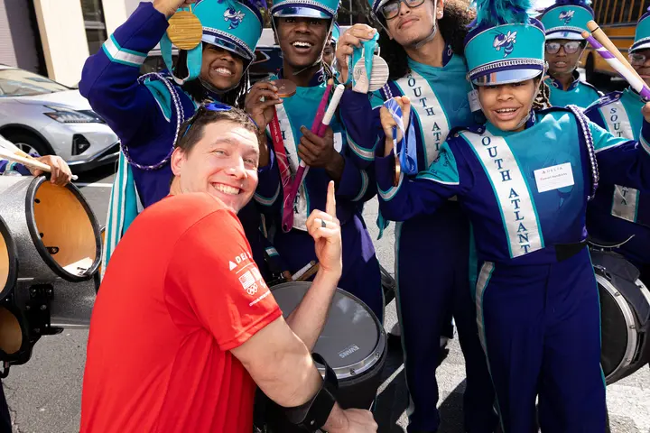 Delta volunteer in a red shirt interacts with a marching band in teal and purple uniforms, with drums and feathered hats, during an outdoor community event.