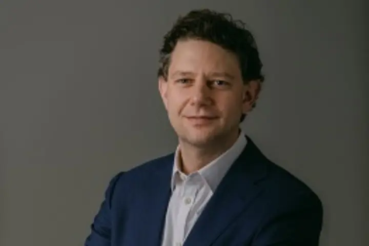 Studio portrait of a person in a dark suit and white shirt, standing with arms crossed against a plain gray background.