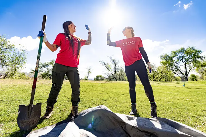 Two Delta volunteers wearing red shirts stand outdoors in a grassy area, raising gloved hands in a high‑five beside a shovel and newly planted tree under a bright sky.