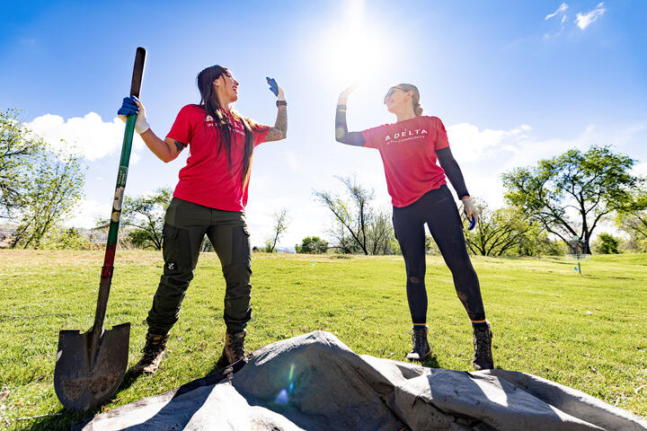 Two Delta volunteers wearing red shirts stand outdoors in a grassy area, raising gloved hands in a high‑five beside a shovel and newly planted tree under a bright sky.