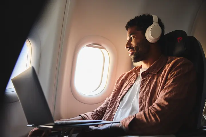 Delta customer wearing headphones works on a laptop while seated by an airplane window, with soft daylight illuminating the aircraft cabin.