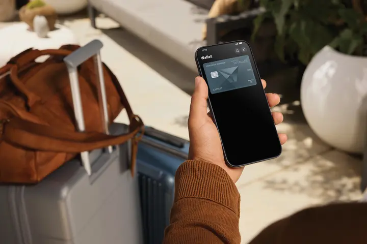 Person holding a smartphone displaying a Delta SkyMiles American Express Card in a digital wallet, with a suitcase and travel bag nearby in a lounge setting.
