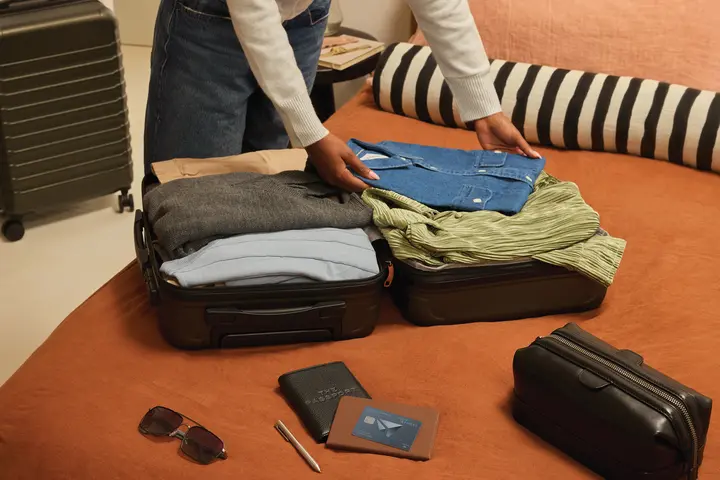 Person packing clothes into an open suitcase on a bed, with a Delta SkyMiles American Express Card, wallet, passport, and sunglasses placed nearby.