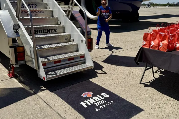 Orange New York Knicks foam finger reading “#1 FAN” sits atop Delta-branded blue towels at an airport display, with travelers and check-in counters in the background.