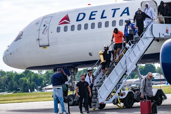 People exit a Delta airplane via stairs on the tarmac, with one person carrying a gold trophy while others hold bags nearby.