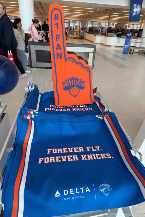 Bright orange New York Knicks foam finger displayed on a stand of blue Delta‑branded rally towels reading “Forever Fly. Forever Knicks” in an airport terminal, with travelers and security area in the background.
