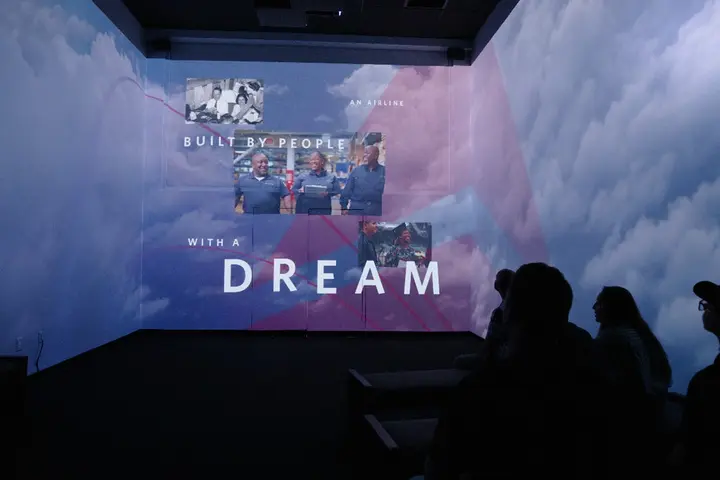 Silhouetted viewers watch an immersive projection of clouds with photos of people and the text “BUILT BY PEOPLE WITH A DREAM.”