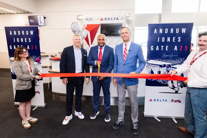 Five people pose at a Delta airport gate ceremony, with the center person holding large scissors to cut a red ribbon. Banners on both sides read “Andruw Jones Gate A25,” featuring baseball-themed artwork and Delta branding.