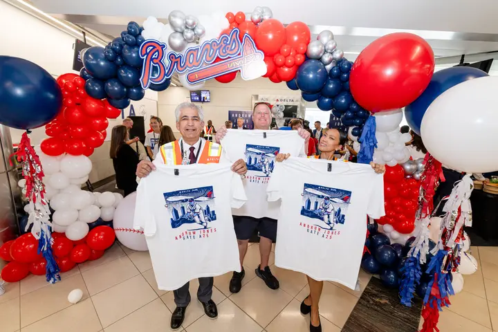 Three attendees stand beneath a Braves-themed balloon arch, displaying commemorative Andruw Jones Gate A25 T-shirts at a Delta airport celebration.