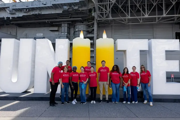 Delta volunteers in red shirts stand in front of a large “UNITE” display with two candle-shaped letters, commemorating 9/11 aboard an aircraft carrier.