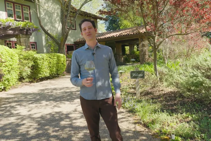 Person holding a glass of white wine on a garden path outside a vineyard-style building surrounded by trees and landscaping.