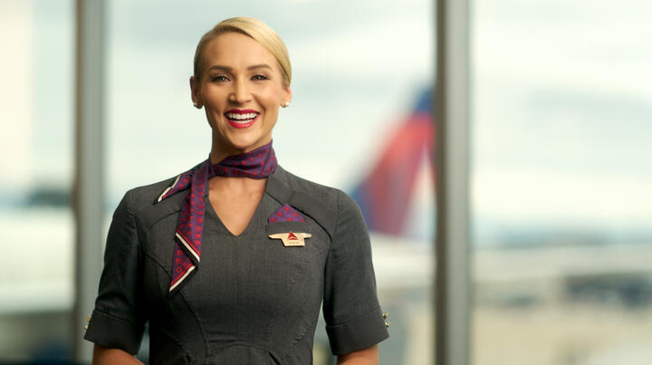 Delta flight attendant smiling, wearing gray dress and purple neck scarf.