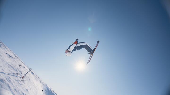 A skier performs a high aerial trick against a clear blue sky on a snowy mountain slope
