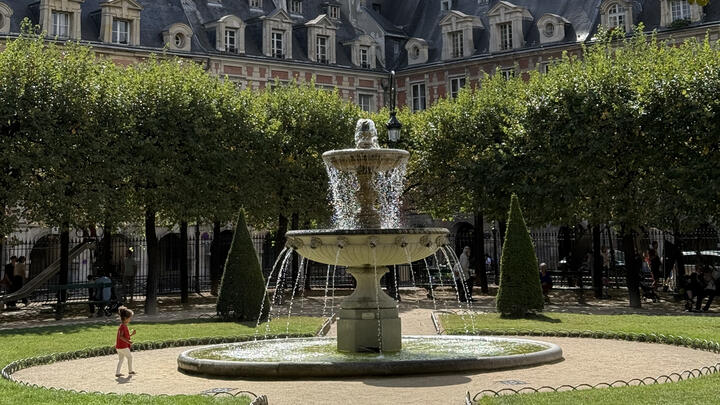 Stone fountain in a leafy Paris square, framed by manicured trees and historic red-brick buildings with slate roofs.