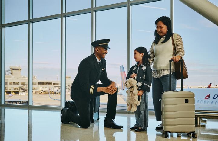 Delta pilot kneeling beside a young child holding a stuffed animal at an airport gate, while an adult stands nearby with rolling luggage; airplane visible through large windows in the background.